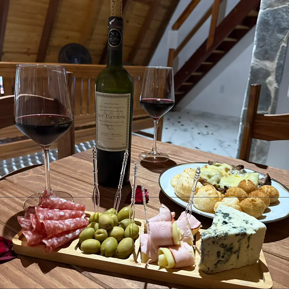 Mesa da sala com vinho, taças e tábua de frios, e a escada para o mezanino ao fundo